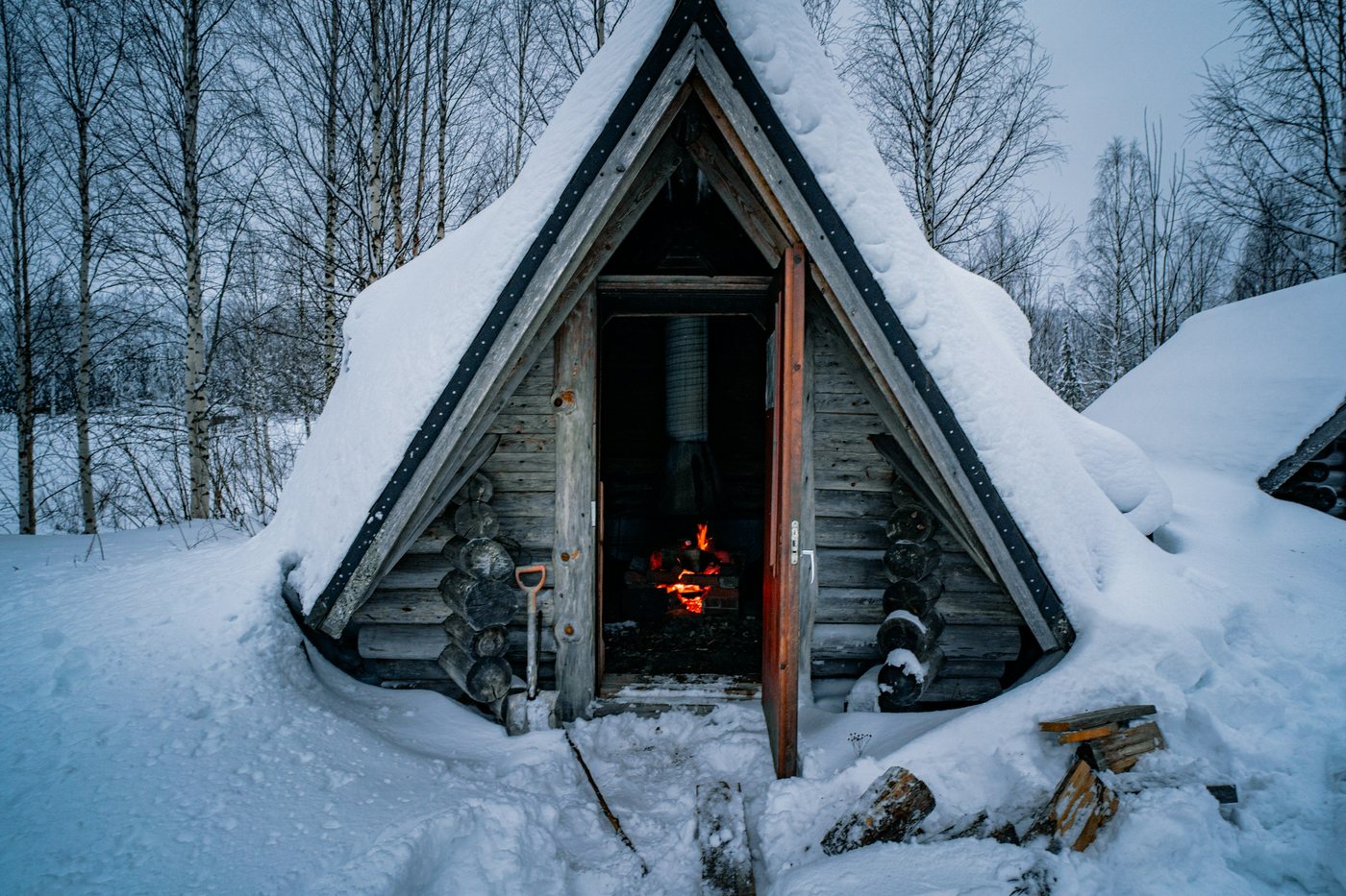 Pequeña cabaña de madera con techo de nieve y un fuego encendido adentro, en un bosque de Laponia